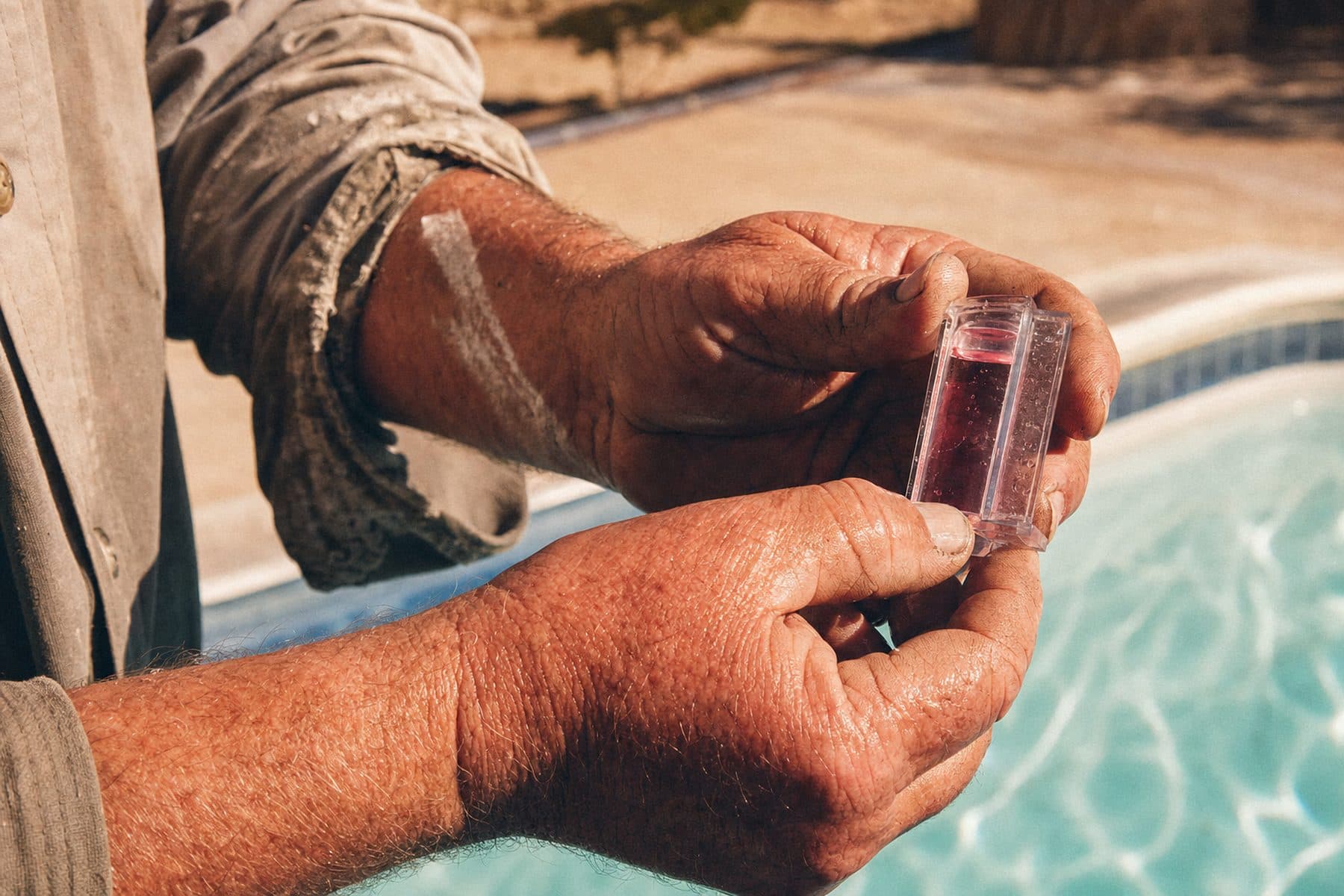 A pool technician's weathered hands holding a water test kit vial of pink reagent water above a turquoise pool in harsh afternoon sun.
